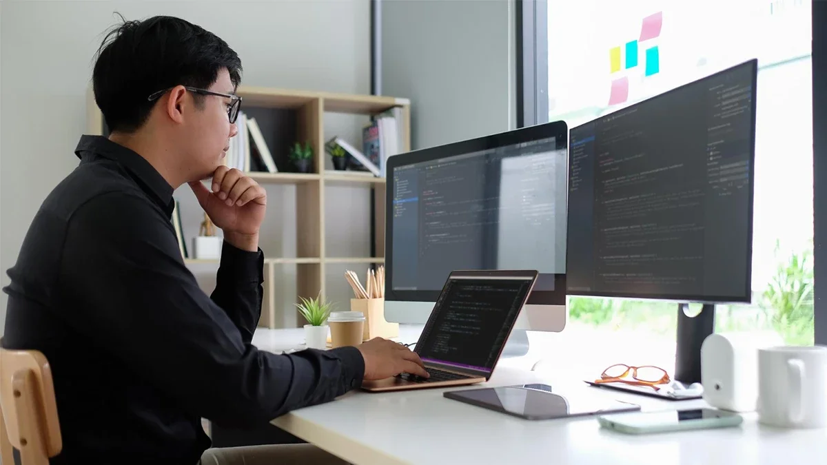 A focused full-stack web developer sitting at a desk with a laptop and two large monitors, working on code in a professional office environment.